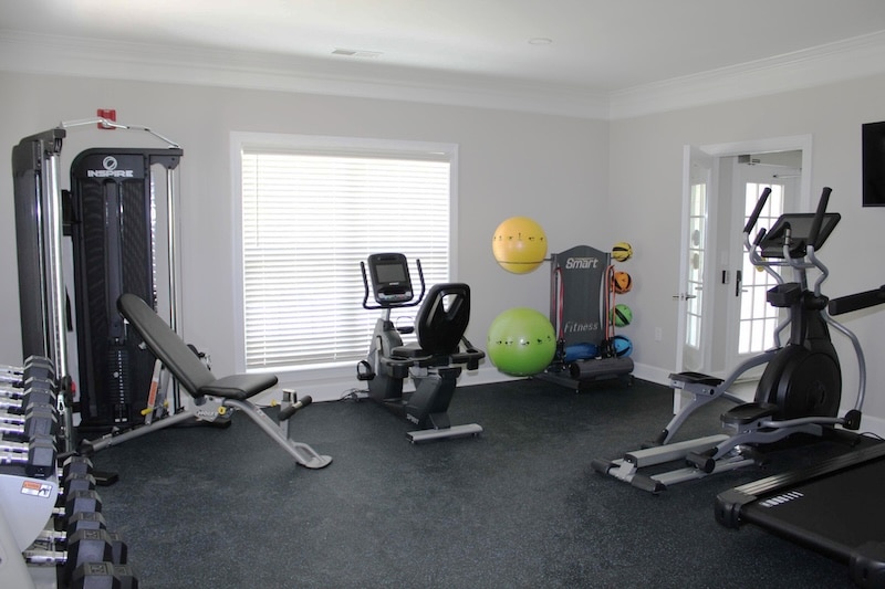 A home gym room with weight machines, a recumbent bike, an elliptical trainer, dumbbells, and several exercise balls near a large window.